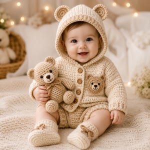 May include: A smiling baby wearing a beige crocheted bear-themed outfit, including a hooded jacket with bear ears, shorts, and booties. The baby holds a matching teddy bear. The outfit has a bear face patch on the chest.