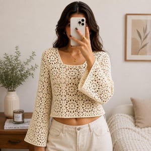 Könnte beinhalten: Cremefarbenes Häkeltop mit quadratischem Ausschnitt und ausgestellten Ärmeln. Das Oberteil hat ein Blumenmuster und wird mit hellfarbigen Jeans kombiniert. Das Bild zeigt eine Person, die ein Telefon vor ihrem Gesicht hält.