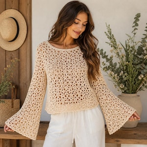 Puede incluir: Un suéter de crochet color crema con tejido abierto y mangas largas acampanadas. El suéter tiene un cuello redondo y se combina con pantalones blancos. Un sombrero de paja cuelga en una pared de madera en el fondo.