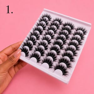 May include: A white plastic tray containing twenty pairs of black false eyelashes arranged in five rows of four. Each pair of lashes features a dense, layered, and wispy design with a curved band. The number 1 is printed in the top left corner.