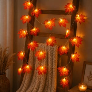 Könnte beinhalten: Eine LED-Lichterkette mit künstlichen Ahornblättern in Orange-, Rot- und Gelbtönen. Jedes Blatt wird von innen beleuchtet und erzeugt einen warmen Schein. Die Lichterkette ist über einen hölzernen Leiterrahmen drapiert.