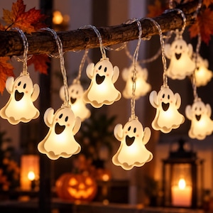 May include: A string of LED lights featuring multiple white, translucent ghost-shaped covers. Each ghost has a black oval mouth, two black eyes, and small pink cheeks. The lights are suspended from a dark, textured branch with autumn leaves.