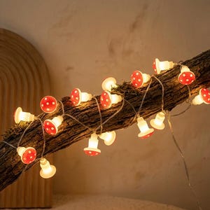 May include: A string of decorative lights featuring small, red mushroom-shaped caps with white polka dots. Each mushroom cap is illuminated from within by a warm white light, connected by a thin, flexible wire draped along a textured tree branch.
