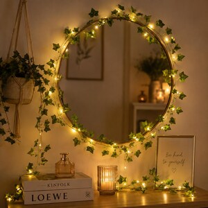May include: A round mirror framed with a string of warm white LED lights and artificial green ivy leaves. The mirror reflects a room interior. Below, a glass candle holder, a perfume bottle, and a framed sign reading Be kind to yourself.