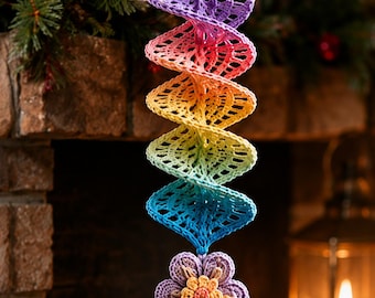 Op de afbeelding: Een hangend gehaakt ornament met een spiraalvormig ontwerp in een regenboogverloop van paars, roze, oranje, geel, groen en blauw. De spiraal is bekroond met drie kleine houten kralen en eindigt met een gehaakte bloem en een veelkleurige kwast.