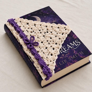 May include: A triangular crochet bookmark made of cream-colored yarn with a purple border and a small purple flower accent. The bookmark is placed on a dark purple book cover with the visible text DREAMS and the author name ELIE SMITH.