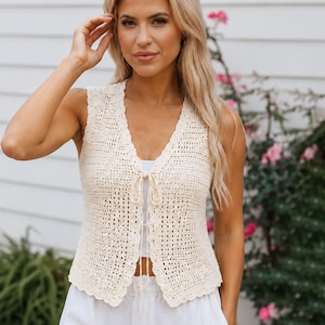 May include: A sleeveless cream-colored crochet vest featuring an open front design with a delicate tie closure at the center. The garment has a scalloped edge along the neckline and hem, showcasing an intricate, airy, and open-knit pattern throughout.