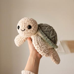 May include: A handmade crochet turtle plushie in cream, gray, and light green. The turtle has a smiling face with black eyes. The plushie is soft and cuddly, perfect for a child's toy or nursery decor.