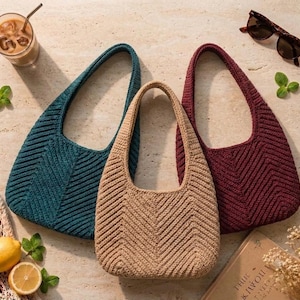 May include: Three crochet hobo bags in teal, beige, and burgundy, each featuring a distinct chevron stitch pattern, a single integrated shoulder strap, and a rounded base. The bags are arranged together, showcasing their textured yarn construction.