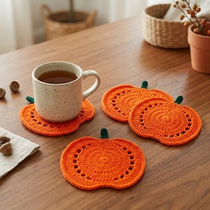 May include: Three orange crochet coasters shaped like pumpkins with small green stems. One coaster supports a speckled beige ceramic mug filled with tea. The coasters feature a circular stitch pattern with decorative openwork holes along the edges.
