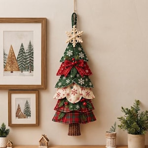 Könnte beinhalten: Ein hängender Weihnachtsbaumschmuck aus Stoff mit geschichteten dreieckigen Paneelen in Grün, Creme und rotem Schottenkaro. Der Baum ist mit weißen Schneeflockenmustern, einer hölzernen Schneeflocke als Spitze, roten Beeren und einem braunen Stoffstamm verziert.