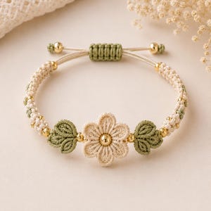 May include: A macrame bracelet featuring a central flower and two leaf accents in beige and green. The bracelet has a braided design with gold-colored beads and an adjustable closure. The flower has a gold-colored center.
