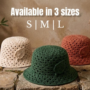 Puede incluir: Tres sombreros de pescador de ganchillo en color crema, verde oscuro y terracota, cada uno con una pequeña aplicación de hoja. El texto "Available in 3 sizes S | M | L" aparece sobre los sombreros, dispuestos en una superficie de piedra.
