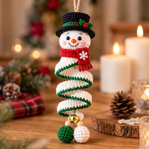 Puede incluir: Un adorno de muñeco de nieve tejido a ganchillo con un sombrero de copa negro con adornos verdes y bayas rojas, una bufanda roja con un copo de nieve blanco y un cuerpo en espiral blanco. El adorno cuelga de un cordel con una campana dorada.