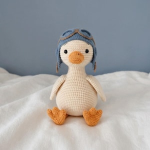 May include: A crocheted duck toy featuring a cream-colored body, bright orange beak and feet, and black bead eyes. The duck wears a blue aviator-style cap with brown trim and goggles, crafted with detailed yarn stitching throughout the entire figure.