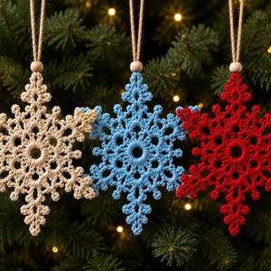 May include: Three crocheted snowflake ornaments in beige, light blue, and red. Each snowflake features a central circular opening, intricate branching patterns, and a small wooden bead at the top, suspended by a thin brown twine loop.