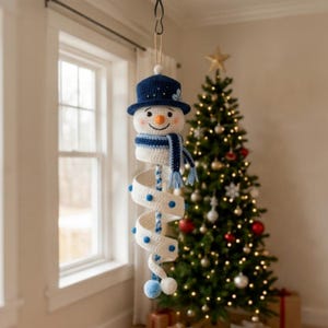 Peut inclure: Une décoration de bonhomme de neige en crochet suspendue, dotée d'un chapeau bleu foncé avec de petits accents blancs, d'un visage souriant avec un nez orange et d'une écharpe rayée bleue et blanche. Le corps est une longue spirale blanche.