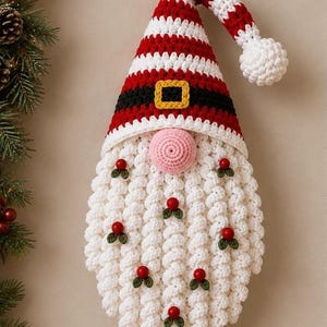 Peut inclure: Une décoration murale en crochet représentant un gnome avec un bonnet rayé rouge et blanc terminé par un pompon, une bande noire à boucle jaune, un nez rose et une longue barbe blanche ornée de petites baies rouges et de feuilles vertes.