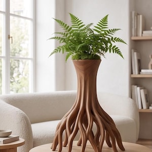 Op de afbeelding: Een bruine houten vaas uit The Arboretum Collection, gevormd als een boom met wortels, bevat een groene varen. De vaas staat op een ronde houten tafel. Op de achtergrond een witte bank en een raam.