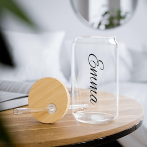 Puede incluir: Un vaso transparente en forma de lata con el nombre "Emma" en escritura negra. Una tapa de bambú natural y una pajita transparente están al lado del vaso. El vaso está sobre una mesa de madera.