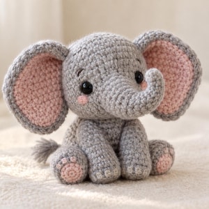 Può includere: Un elefante di peluche lavorato all'uncinetto di colore grigio con grandi orecchie che presentano centri rosa chiaro. L'elefante ha occhi di sicurezza neri, piccoli segni di rossore rosa sulle guance e cuscinetti circolari rosa chiaro.