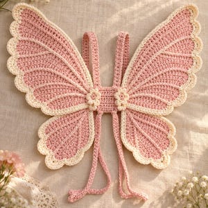 May include: A pair of handcrafted crochet butterfly wings in soft pink with a cream-colored scalloped border. The wings feature intricate stitch detailing and two small flower accents at the center, with two long pink straps attached for wearing.