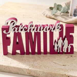 Könnte beinhalten: Ein magenta-farbenes Holzschild mit dem weißen Schriftzug "Patchwork FAMILY". Das Wort "FAMILY" enthält eine weiße Silhouette einer Familie. Ein dekorativer Artikel für die Inneneinrichtung.