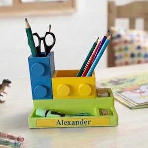 Könnte beinhalten: Ein mehrfarbiger Schreibtisch-Organizer in Form von Bausteinen mit einem blauen vertikalen Abschnitt, einem gelben quadratischen Fach und einer hellgrünen Basis. Das gelbe Etikett vorne zeigt den Namen Alexander in schwarzer Schrift.