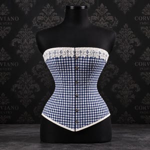 May include: A strapless corset featuring a blue and white gingham check pattern, a white lace trim along the top edge, and a vertical row of metal busk fasteners down the center front. The garment is displayed on a black mannequin torso.
