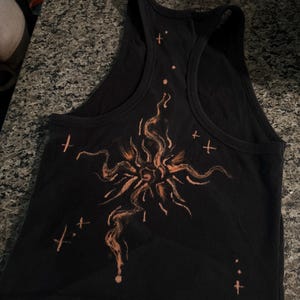 Könnte beinhalten: Ein schwarzes, geripptes Tanktop mit Ringerrücken, das in der Mitte eine orangefarbene Grafik einer stilisierten Sonne mit wellenförmigen Strahlen zeigt. Das Design ist von kleinen orangefarbenen Kreuzen und Punkten umgeben.