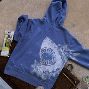 May include: A periwinkle blue hooded sweatshirt featuring a large, hand-painted white shark graphic on the back. The shark is depicted with an open mouth showing sharp teeth, surrounded by stylized water splashes and white paint speckles.