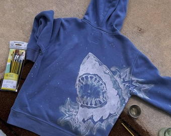 May include: A periwinkle blue hooded sweatshirt featuring a large, hand-painted white shark graphic on the back with an open mouth and sharp teeth. The design includes white paint splatters across the fabric, accompanied by a pack of paintbrushes.