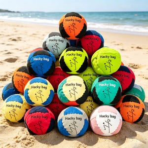 May include: A collection of colorful hacky sacks, each with a unique color scheme and the words "Hacky bag" printed on them. The hacky sacks are arranged in a pile on a sandy beach, with the ocean visible in the background. Each hacky sack features a stylized figure.