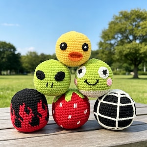Op de afbeelding: Een verzameling van zes bolvormige gehaakte speeltjes met diverse ontwerpen: een geel kuikentje, een groene alien, een groene kikker met roze wangen, een zwarte bal met rode vlammen, een rode aardbei met witte zaadjes en een zwarte bal met witte rasterlijnen.