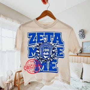 May include: A cream-colored short-sleeve t-shirt featuring a large blue graphic with the text ZETA PHI BETA and the year 1920. The design includes a central shield emblem with a book and dove, surrounded by a shattered stone effect.