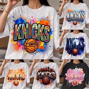 Op de afbeelding: Een wit t-shirt met korte mouwen voorzien van een grote grafische print van het woord KNICKS in dikke letters gevuld met een kleurrijke illustratie van de skyline van New York, vergezeld door een basketbalafbeelding en levendige aquarelverfspatten.