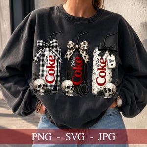 May include: A charcoal gray sweatshirt featuring a graphic of three Diet Coke cans, each topped with a straw and a bow. The cans are decorated with skulls, roses, and patterns including buffalo plaid and skulls. One can has a tag that says Boo.