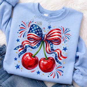 Puede incluir: Sudadera azul claro con un diseño patriótico. El gráfico incluye un lazo con el patrón de la bandera estadounidense, dos cerezas rojas, fuegos artificiales y estrellas. El diseño está centrado en la parte delantera de la sudadera.