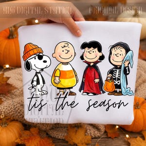 May include: A white sweatshirt featuring a graphic of four cartoon characters dressed in Halloween costumes. Snoopy wears an orange beanie, Charlie Brown wears a candy corn shirt, Lucy wears a vampire cape, and Linus wears a skeleton suit. The text reads 'tis the season' in black cursive script.