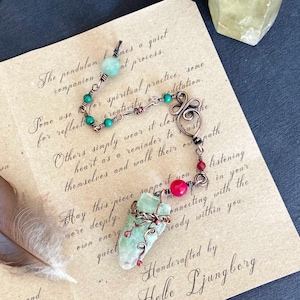 Puede incluir: Un colgante de cristal hecho a mano con detalles de alambre de cobre, cuentas verdes y rojas y un gran cristal verde. El colgante está sobre una nota manuscrita. Otros cristales y una pluma están en el fondo.