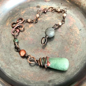 Op de afbeelding: Een handgemaakte koperdraadketting met een groene druppelvormige hanger. De ketting heeft een reeks kleine kralen in verschillende kleuren en een grotere grijze steen. De ketting wordt gepresenteerd op een aangetaste zilverkleurige plaat.