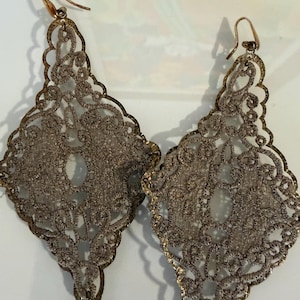 Pode incluir: Um par de brincos pendentes em tom dourado com um design intrincado de filigrana vazada em forma de diamante. Os brincos são construídos com um padrão de metal delicado semelhante a renda e estão presos a ganchos de metal dourado simples.