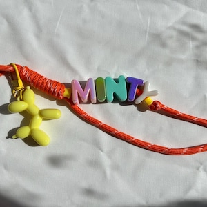 Puede incluir: Un llavero con un cordón naranja brillante, un mosquetón verde azulado, un dije de perro globo amarillo y cuentas de letras de plástico de colores que forman la palabra MINTY. El cordón está envuelto en hilo naranja e incluye cuentas amarillas.