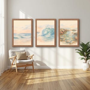 Op de afbeelding: Drie ingelijste wandkunstwerken met oceaanlandschappen in aquarel-stijl. De linker print toont een rustige kustlijn, het midden een grote brekende golf en de rechter rotsachtige getijdenpoelen. Elke print zit in een lichtbruine houten lijst.