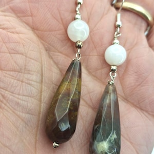 Può includere: Un paio di orecchini pendenti con pietre marroni e verdi sfaccettate a forma di goccia, sospese a ganci in metallo color argento. Ogni orecchino presenta una perla bianca rotonda e liscia posizionata sopra la pietra più grande e scura.
