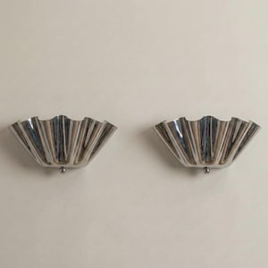 May include: Two matching, silver-colored wall sconces with a scalloped, fan-like design. Each sconce has a small, round detail at the bottom. The sconces are mounted on a plain, off-white wall.