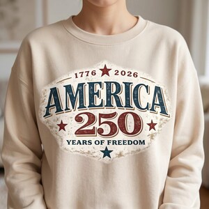 Pu&ograve; includere: Felpa beige con una grafica invecchiata che celebra il 250&deg; anniversario dell'America. Il design presenta la parola "AMERICA" in blu navy, il numero "250" in bordeaux e gli anni "1776" e "2026" con stelle.