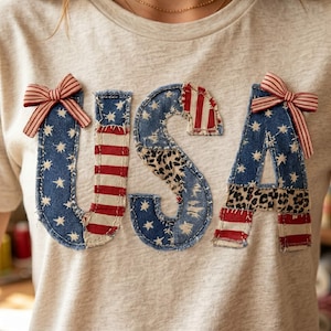 Op de afbeelding: Lichtgrijs t-shirt met het woord "USA" op de voorkant. De letters zijn gemaakt van denim, rood-wit gestreepte stof en luipaardprintstof. Elke letter is versierd met een rood-wit gestreepte strik.