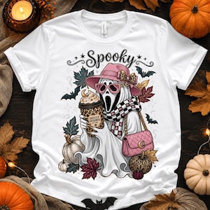 May include: A white t-shirt featuring a graphic of a ghost wearing a pink hat, pink sunglasses, and a checkered scarf. The ghost holds a leopard print coffee cup labeled Ghostly Grounds and carries a pink quilted handbag, surrounded by autumn leaves.