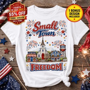 May include: A white t-shirt featuring a graphic of a small town with a church, a water tower, and buildings. The text reads Small Town FREEDOM, USA 240998. The design includes red, white, and blue fireworks, stars, and patriotic accents.